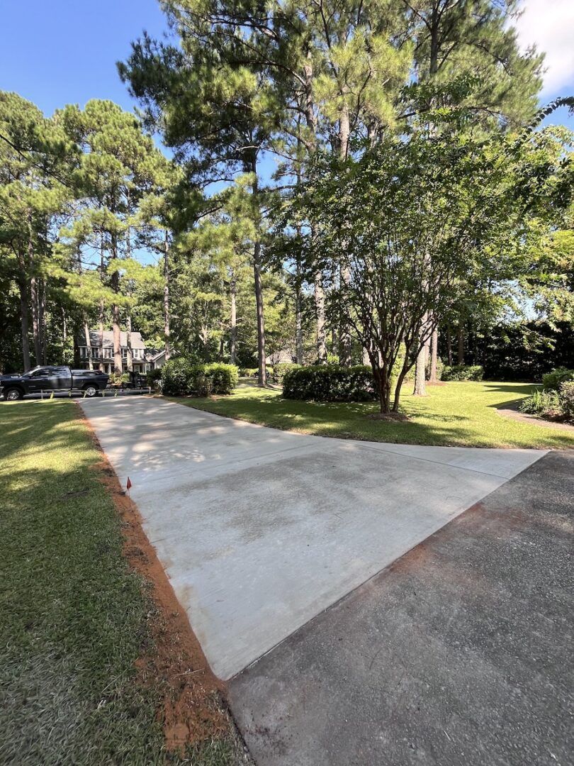 A freshly poured light gray concrete driveway extension sits next to an existing darker asphalt driveway in a wooded yard.