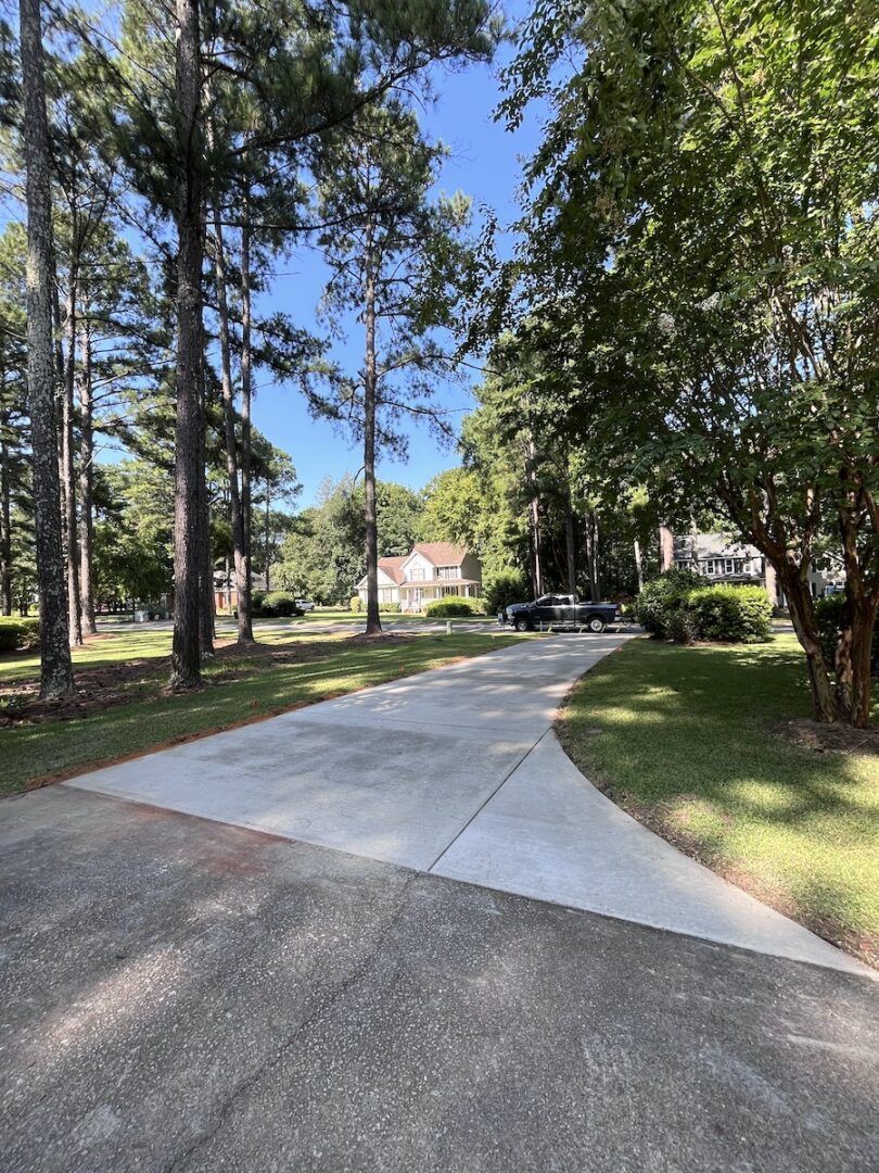 A paved driveway leads toward a two-story house surrounded by tall pine trees and green lawn under a clear blue sky.