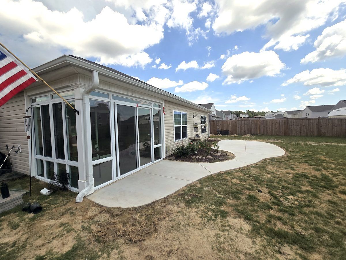 A beige house with a sunroom and a curved concrete patio in the backyard under a blue, cloudy sky.