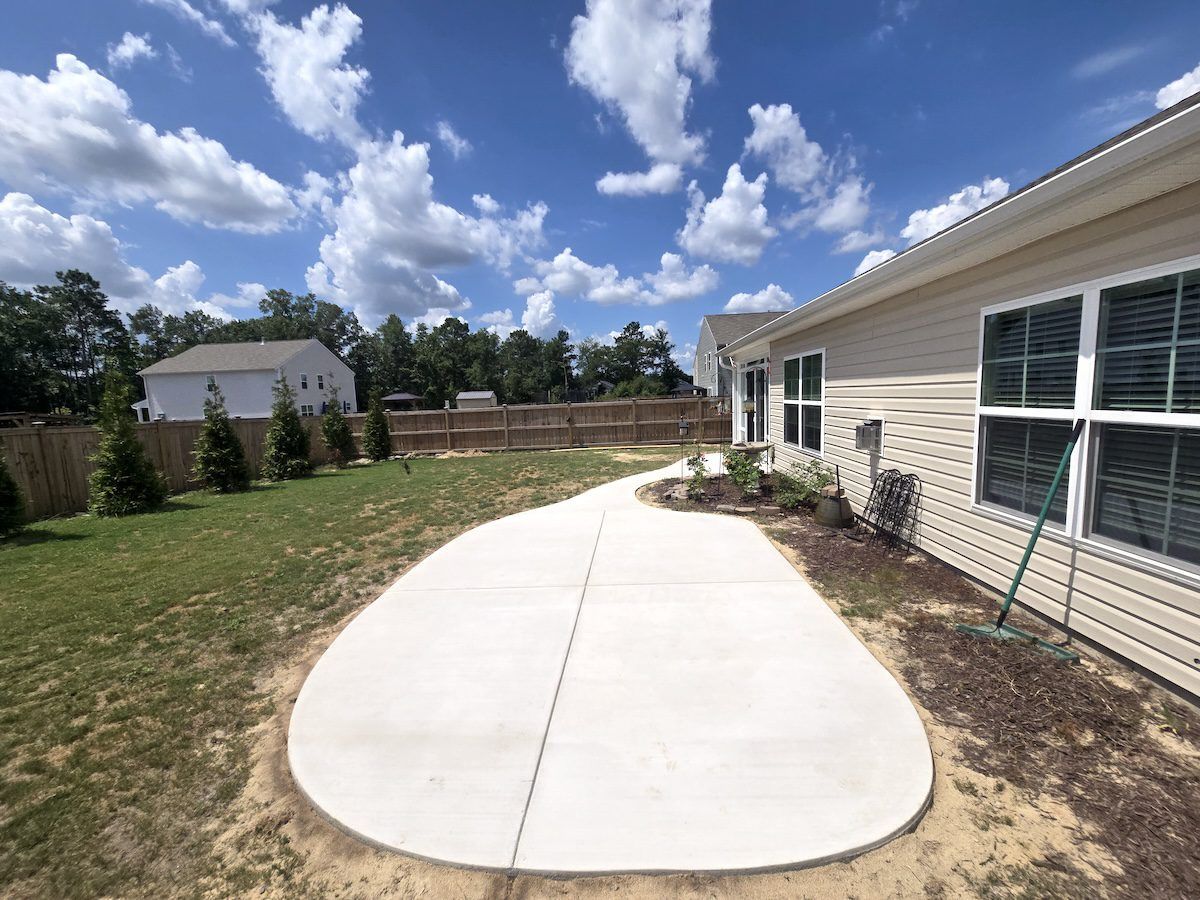 A new concrete patio with a rounded edge extends from the side of a light-colored house into a grassy backyard.