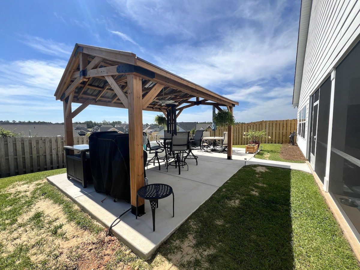 A wooden pergola with a corrugated metal roof stands on a concrete patio in a backyard next to a house.