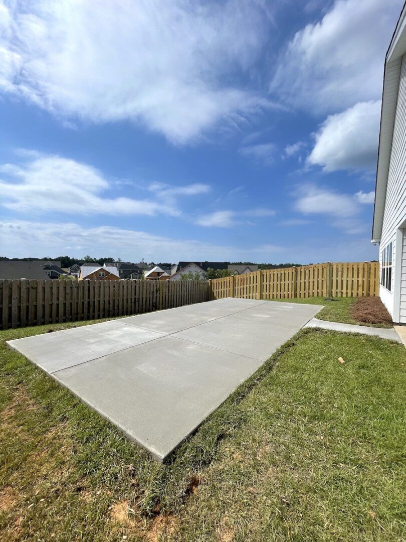 A rectangular concrete patio slab in a backyard with a wooden fence under a blue sky with scattered clouds.