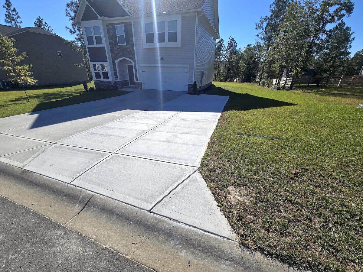 A freshly poured concrete driveway leads to a two-story house on a sunny day with green grass in the surrounding yard.