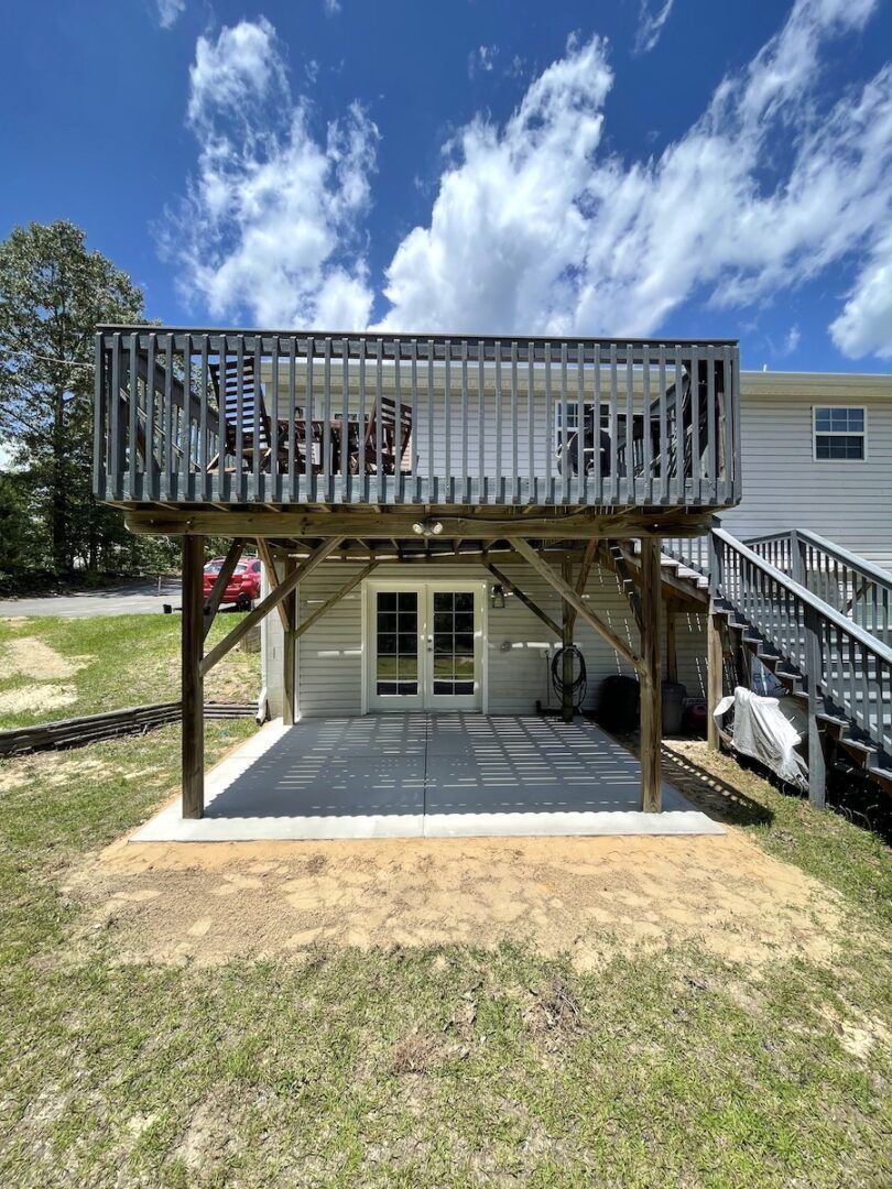 A two-story house exterior featuring a wooden upper deck over a concrete patio with French doors, viewed from the yard.