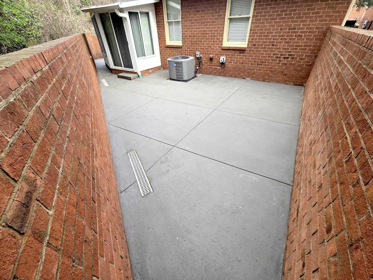A gray concrete patio nestled between two red brick walls, featuring a drainage grate and an outdoor HVAC unit.