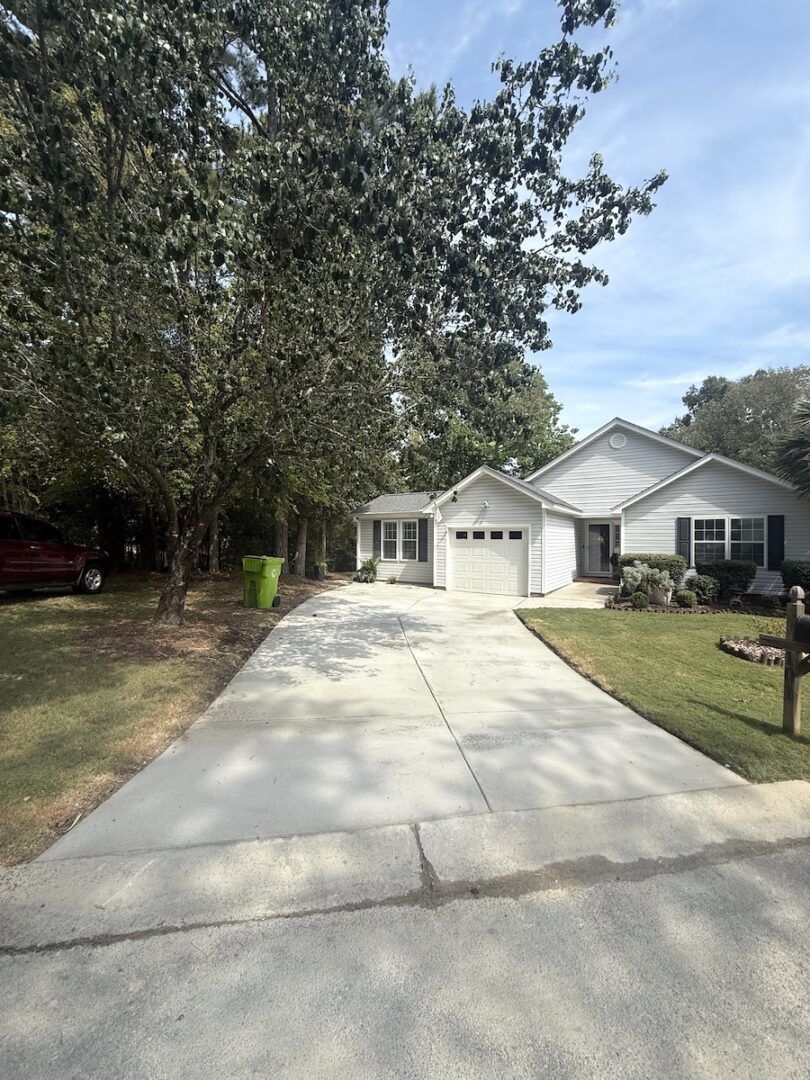 A suburban one-story house with white siding and a concrete driveway leading to a garage under a large, leafy tree.