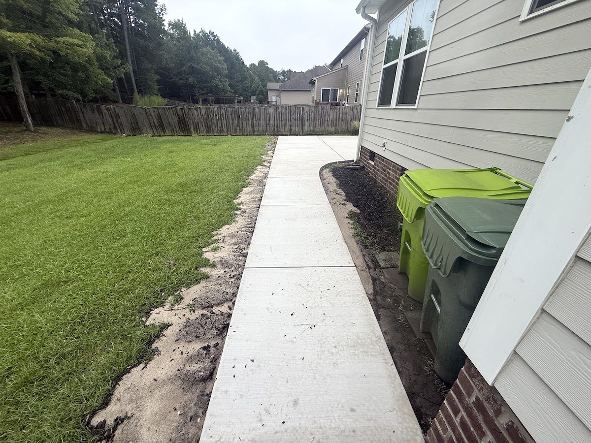 A concrete walkway runs along the side of a house, flanked by a lawn on the left and two trash bins on the right.