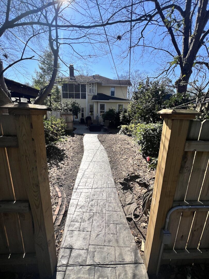A stone walkway leads from a wooden fence gate through a yard toward a two-story beige house on a sunny day.