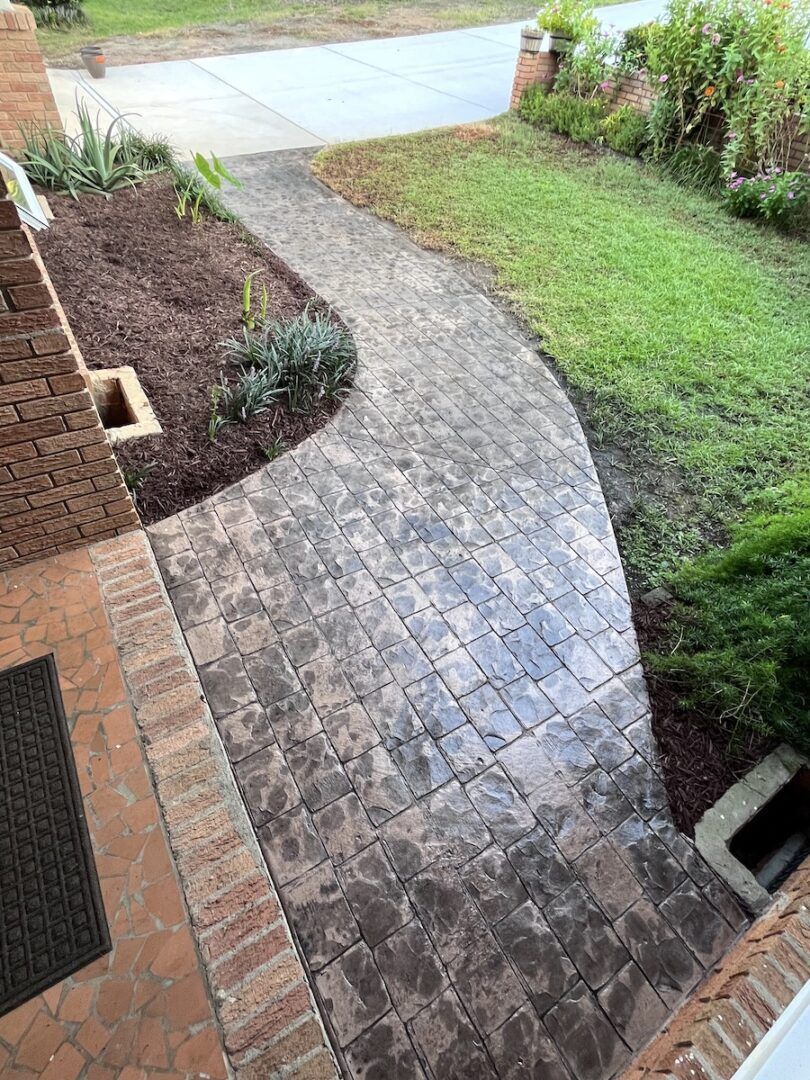 A stamped concrete walkway with a brick border leads from a porch across a grassy lawn to a driveway.