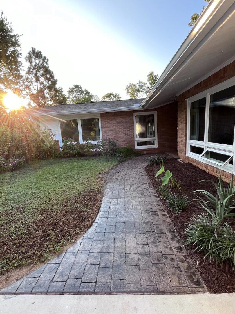 A brick walkway leads to a mid-century brick house at sunset, with greenery in the yard and large white-framed windows.