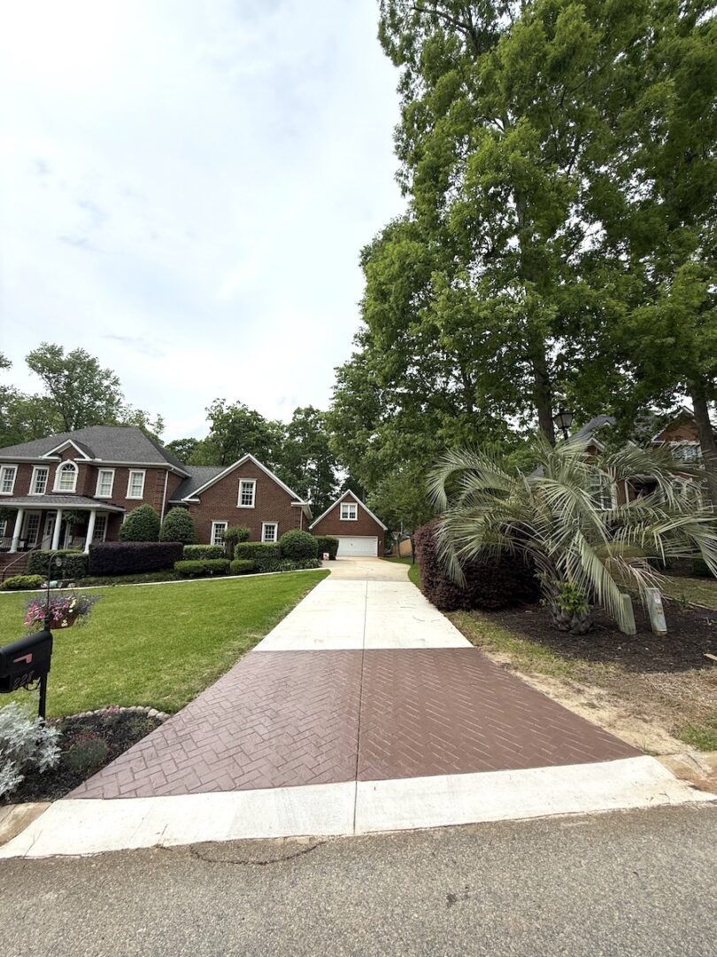 A brick-front house viewed from the street with a long driveway featuring a textured, reddish-brown paved section.