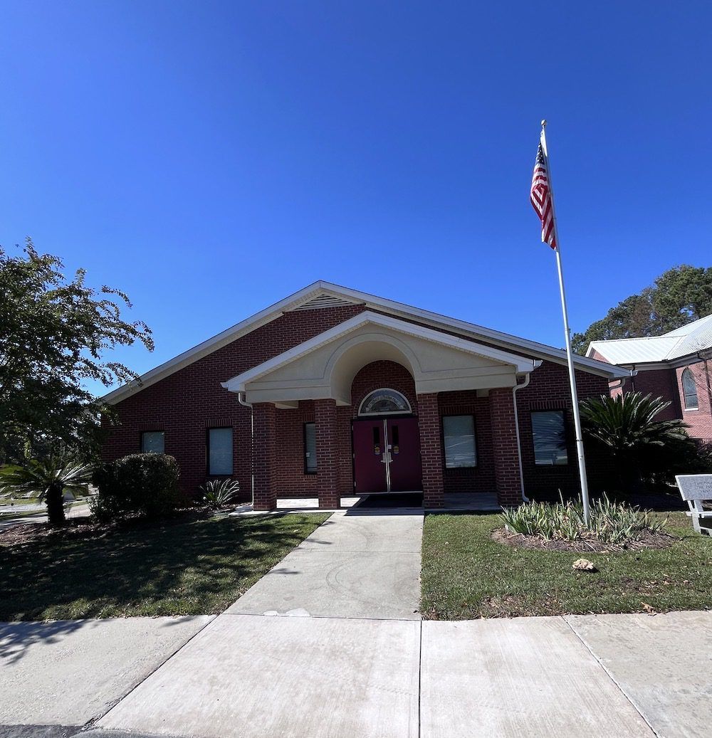 A red brick building with a covered entrance and a tall American flag flying on a pole in the front yard under blue sky.