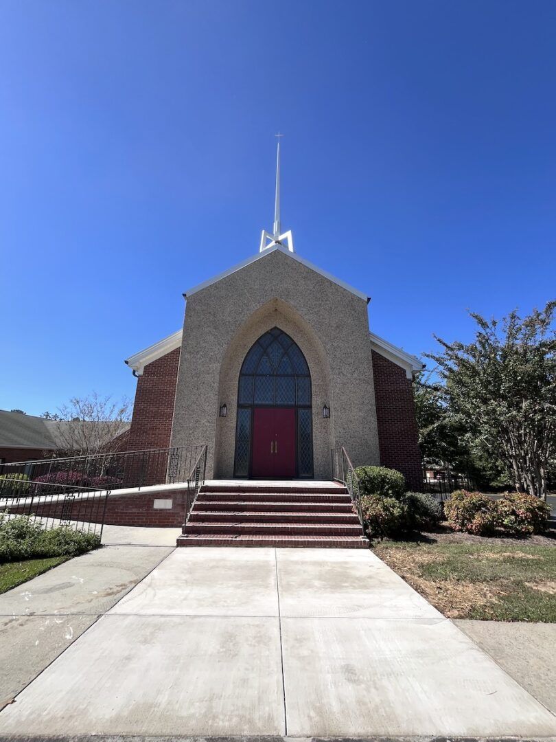 A stone church with a red door and a pointed steeple under a clear blue sky, accessed by a set of concrete steps.