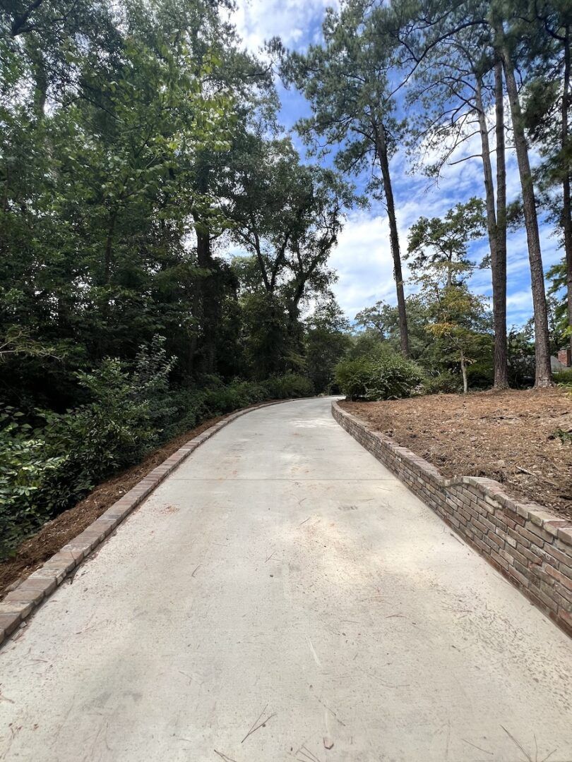 A concrete driveway curves through a wooded area lined with a short stone retaining wall under a blue sky.