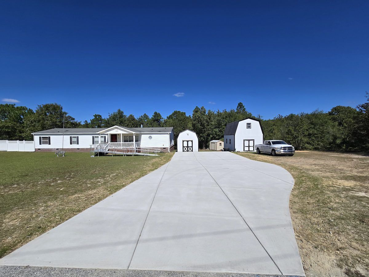 A white, single-story home with a porch, a large concrete driveway, two outbuildings, and a truck under a blue sky.