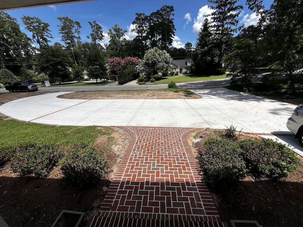 A red brick walkway leads from the foreground toward a newly poured concrete driveway in front of a house.