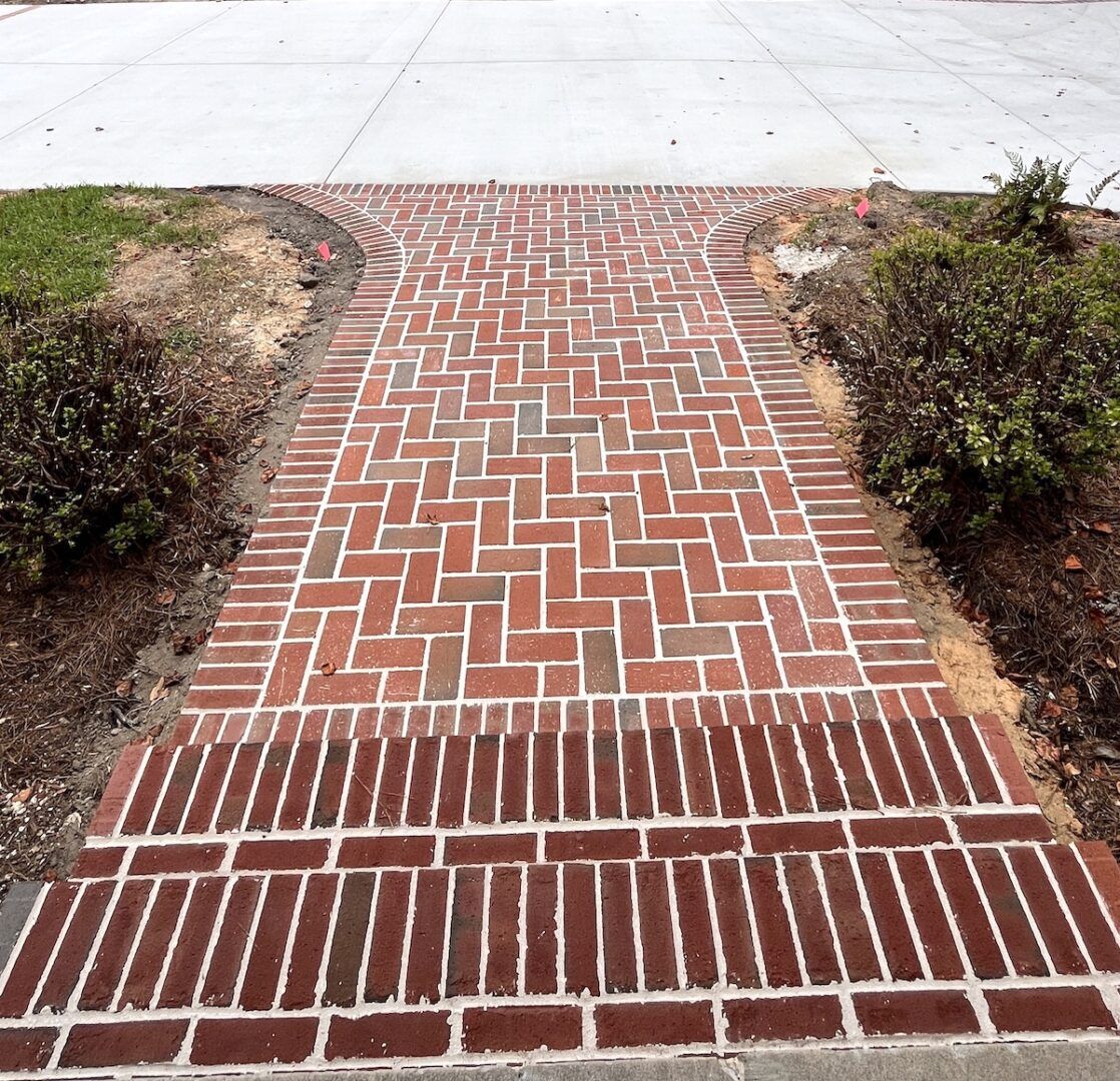 A brick walkway with a herringbone pattern leads to a set of stairs, flanked by garden beds and a concrete driveway.