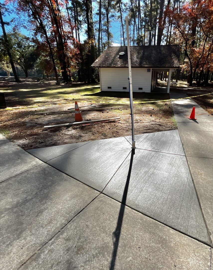 A freshly poured triangular concrete pad surrounds a vertical pole, near a small white building and orange safety cones.