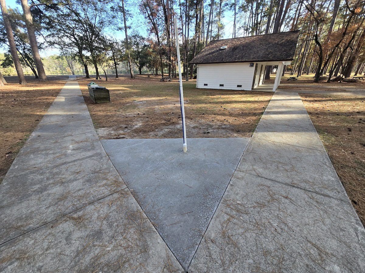 A white building and a flagpole stand beyond a Y-shaped concrete path in a wooded, grassy clearing.