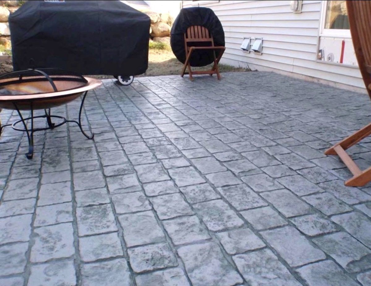 A gray stamped concrete patio with a fire pit, a covered grill, and a wooden chair near a house.