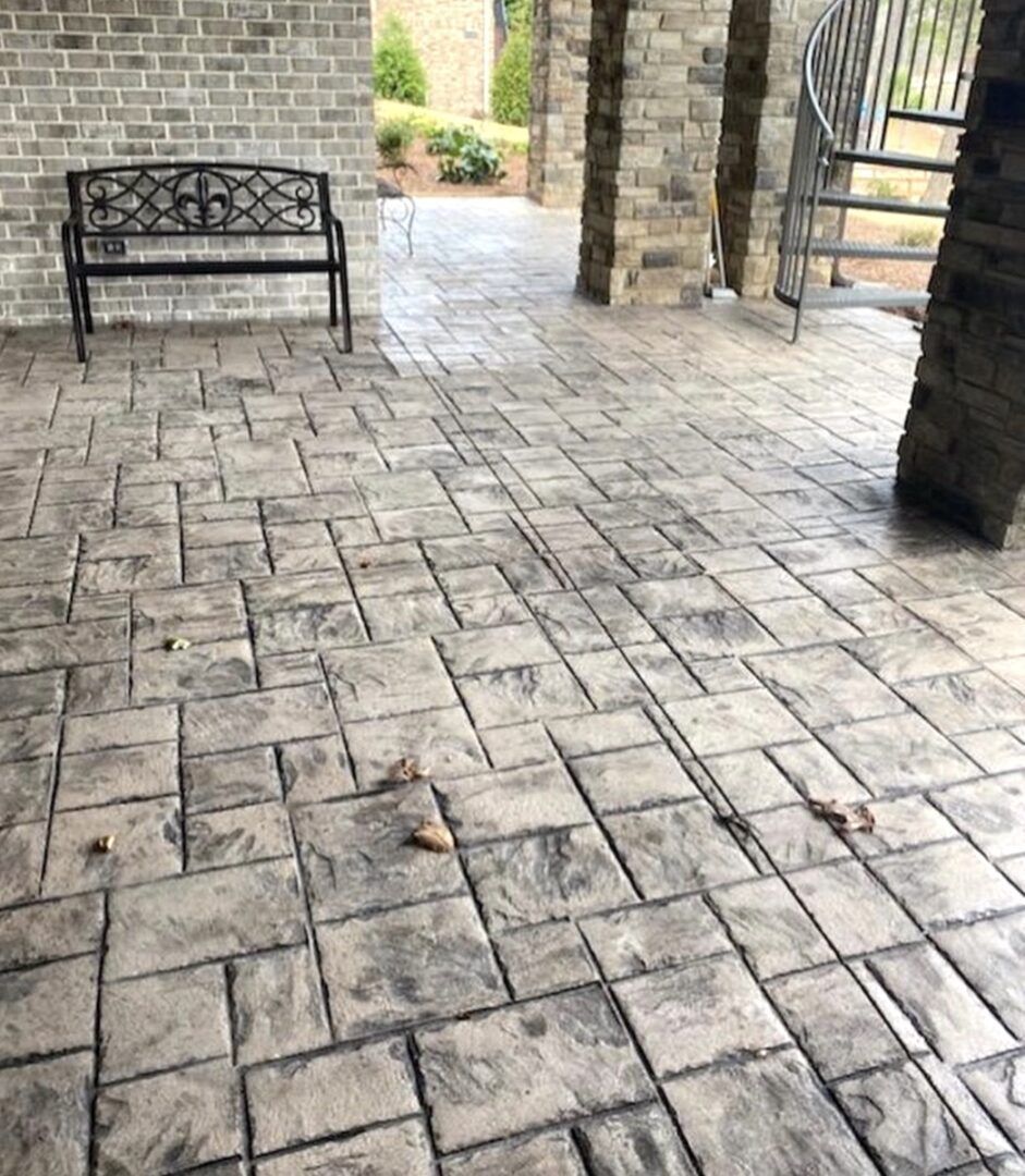 A patio with stone-patterned concrete flooring, a black metal bench, stone pillars, and a spiral staircase outdoors.