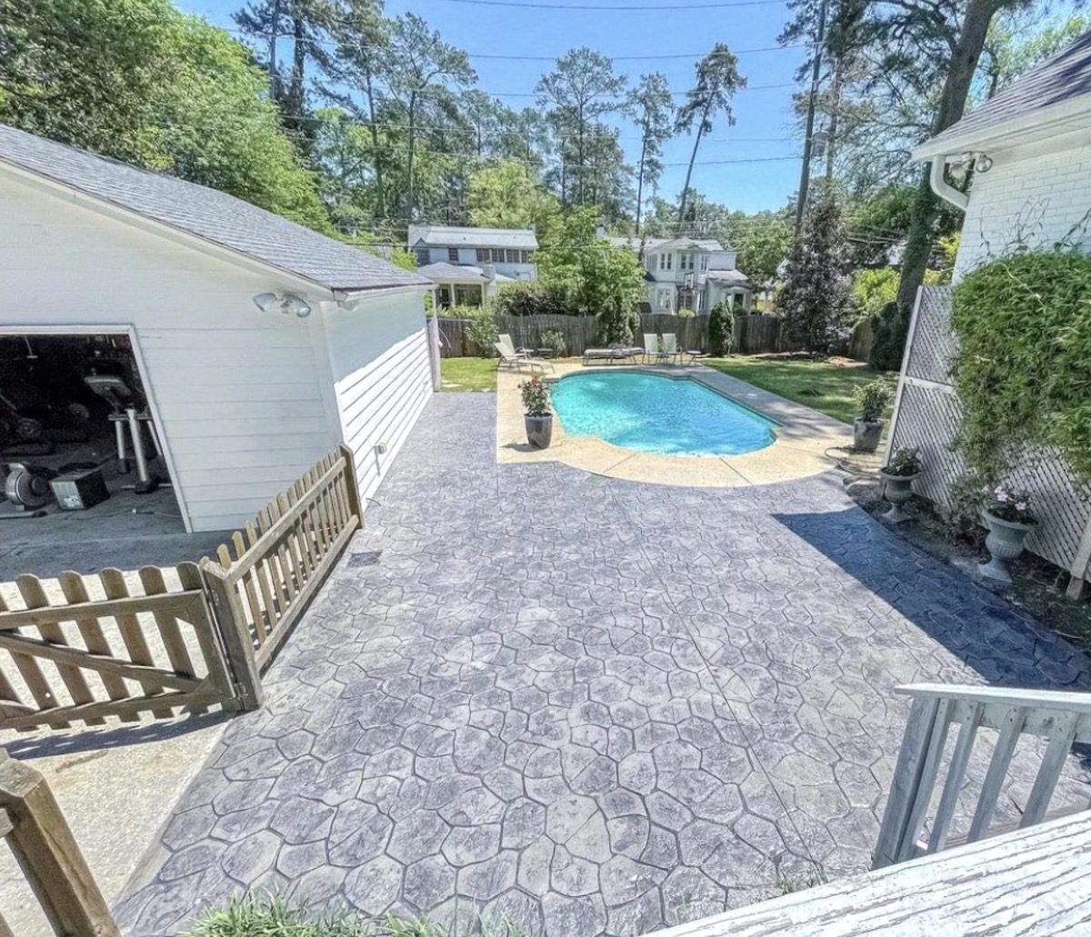 A backyard patio with gray stamped concrete, a small blue pool, a white detached garage, and surrounding trees.