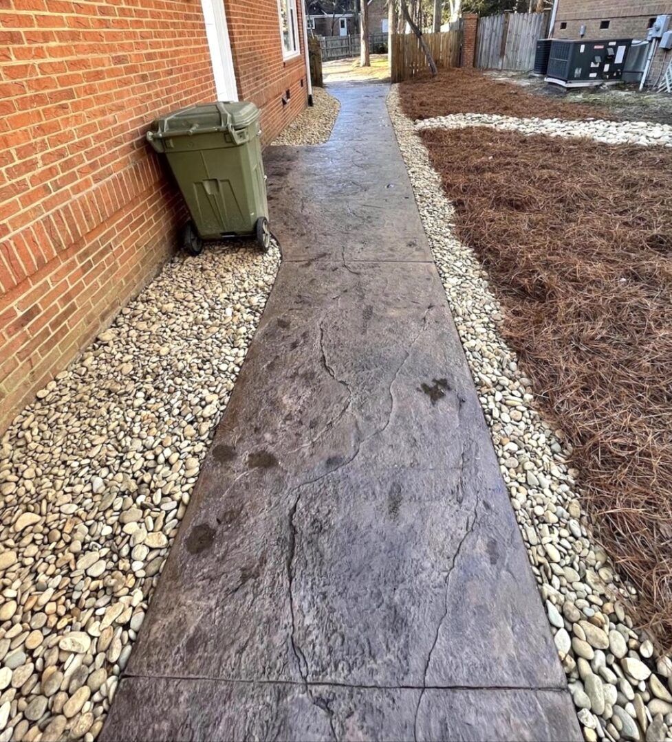 A stamped concrete walkway between a brick wall and a stone bed, with a green trash bin on the left.