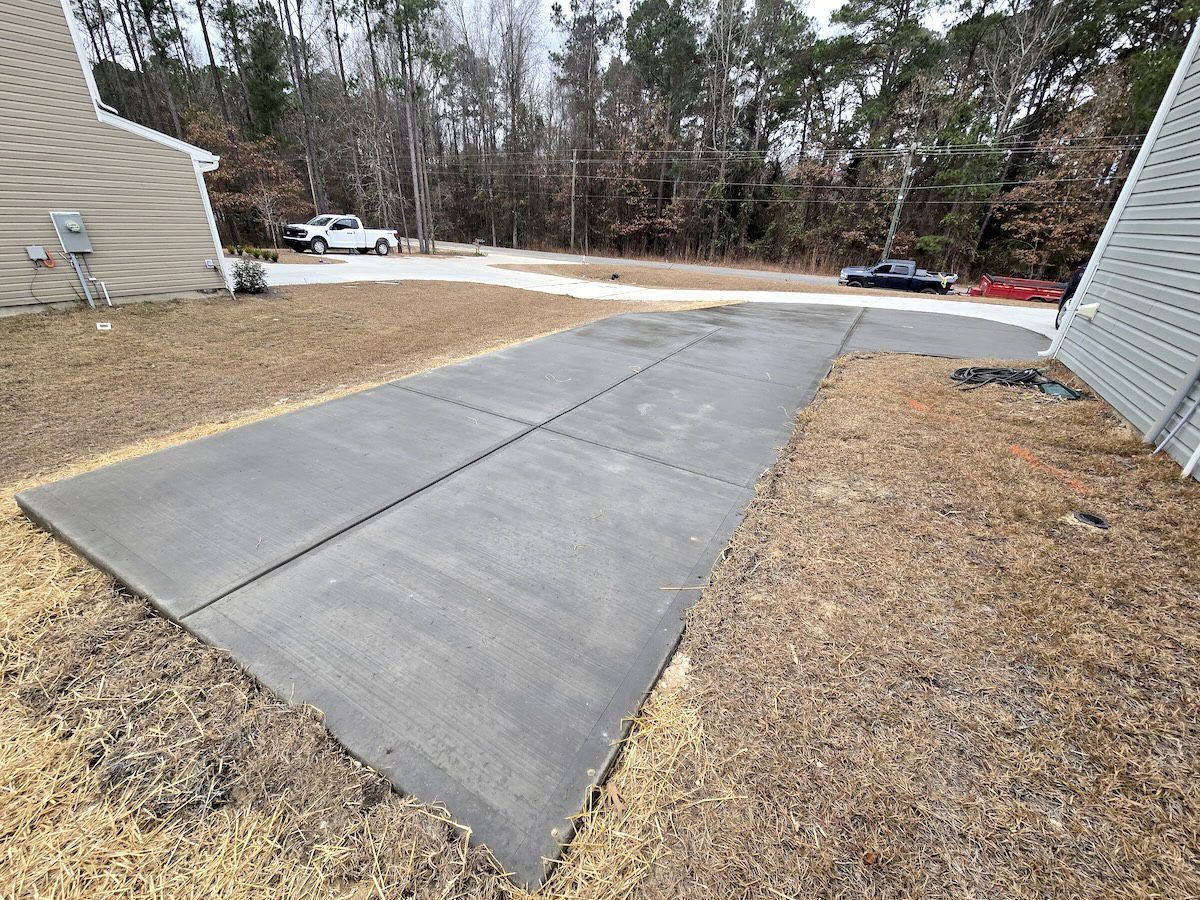 A newly poured concrete driveway extension sits between two houses, surrounded by dormant grass and trees.