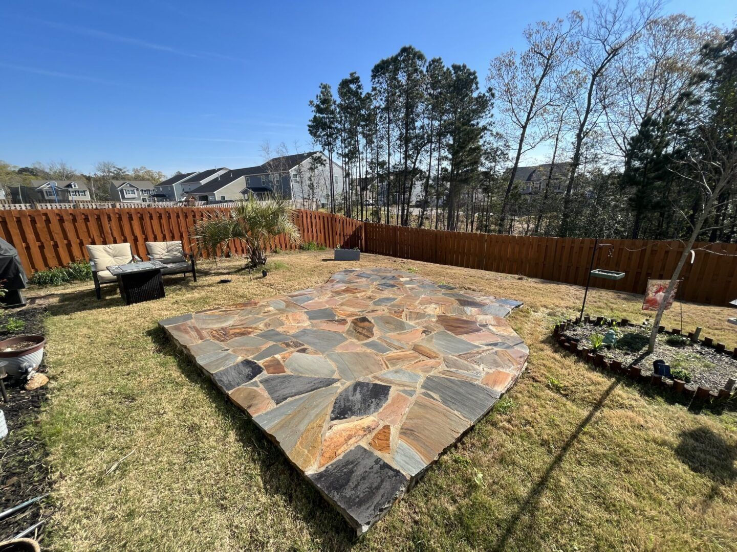 A flagstone patio in a backyard with a brown wooden fence, lawn chairs, and trees under a clear blue sky.