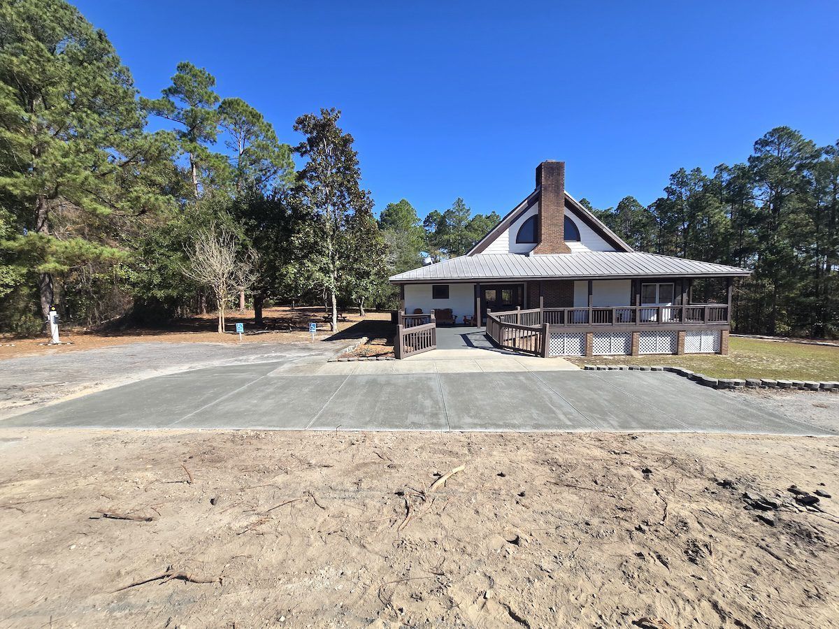 A building with a covered porch and accessible ramp, positioned behind a newly poured concrete driveway in a wooded area.