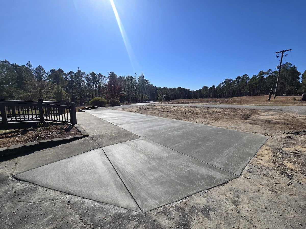 A newly poured concrete driveway leads toward an open, dirt-covered lot on a bright, sunny day with trees in the distance.