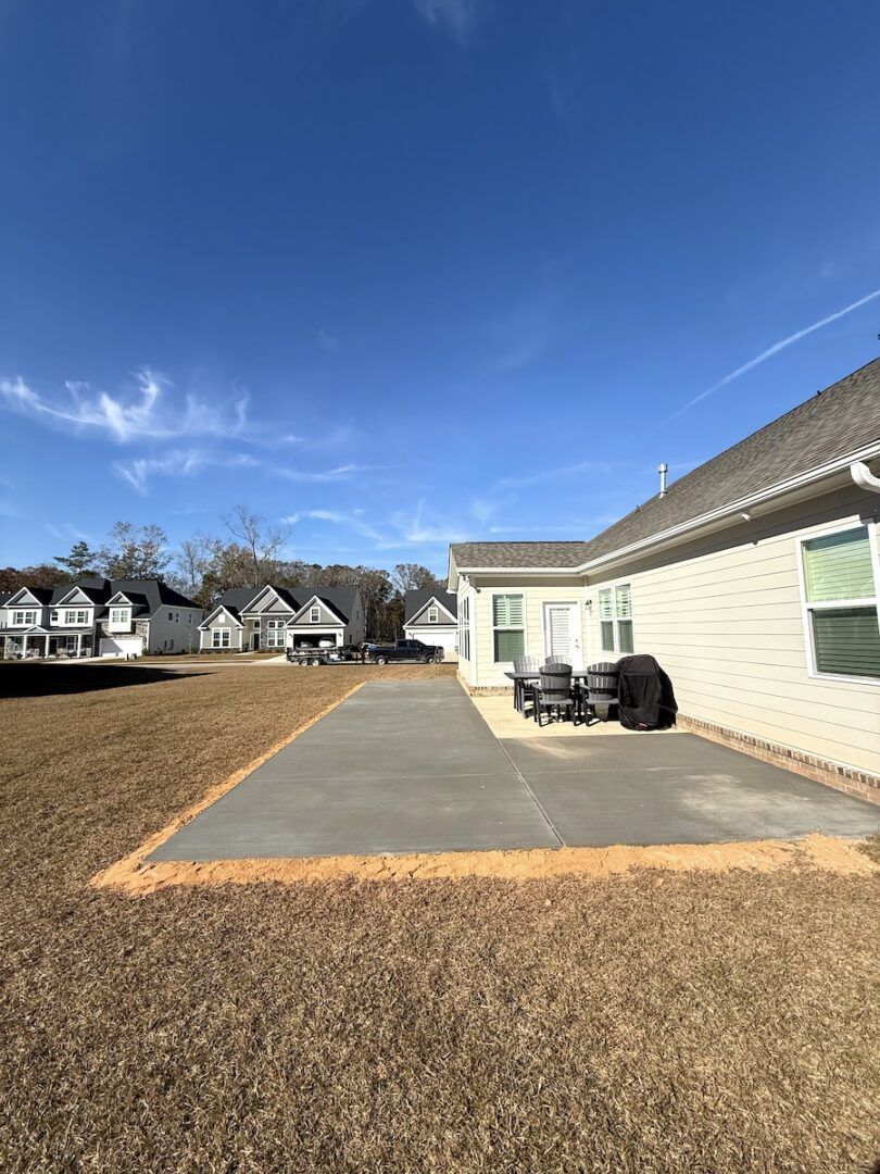 A beige house with a new rectangular concrete patio sits on a grassy lot under a clear blue sky.