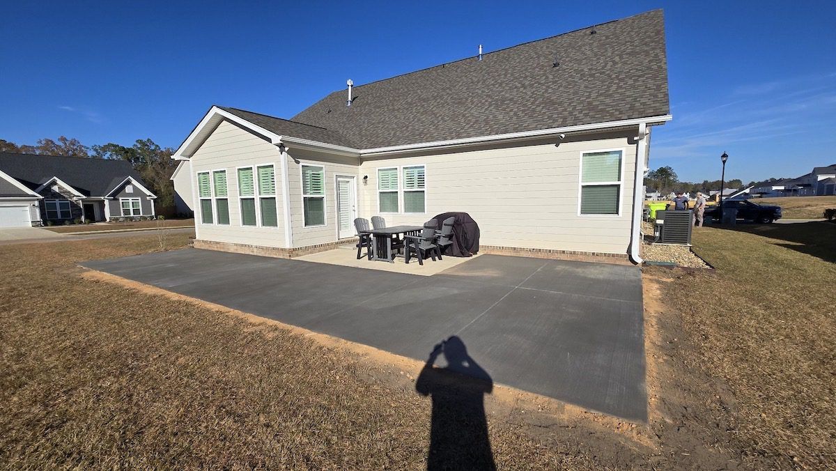 A backyard view of a light-colored house with a large, rectangular concrete patio and a small dining set on a sunny day.