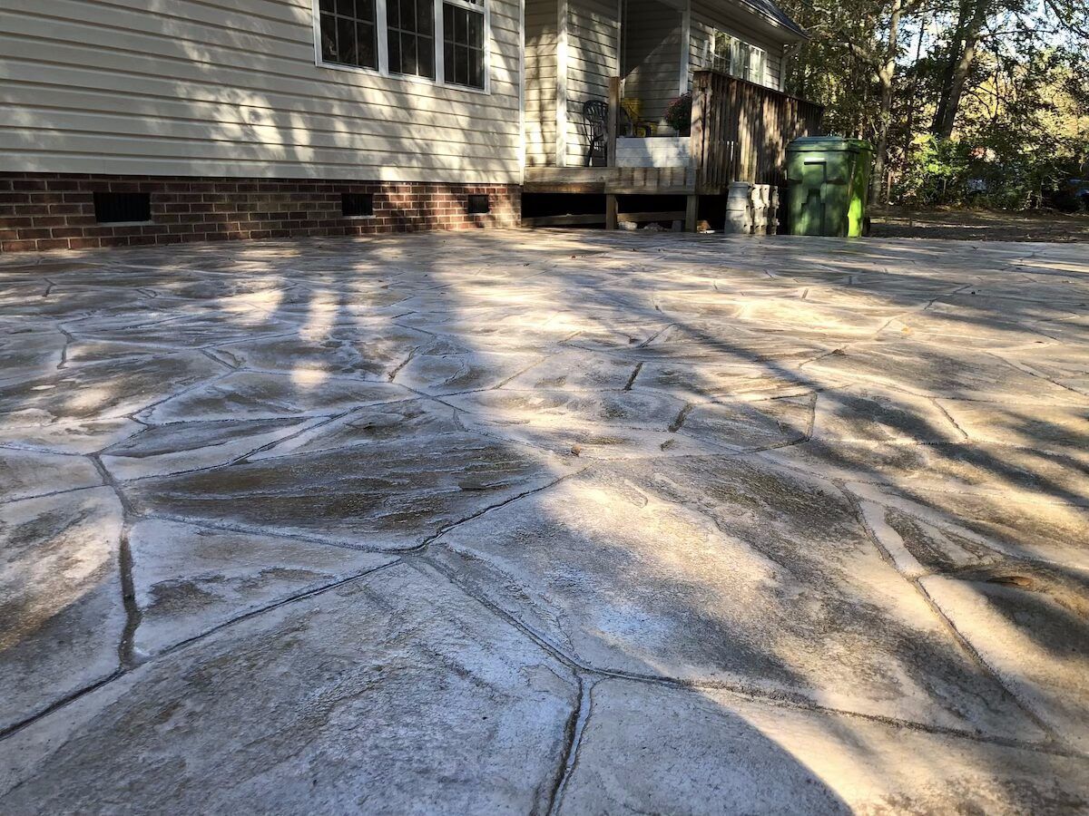 Stamped concrete patio with a stone-like pattern, next to a residential house with siding and a brick foundation.