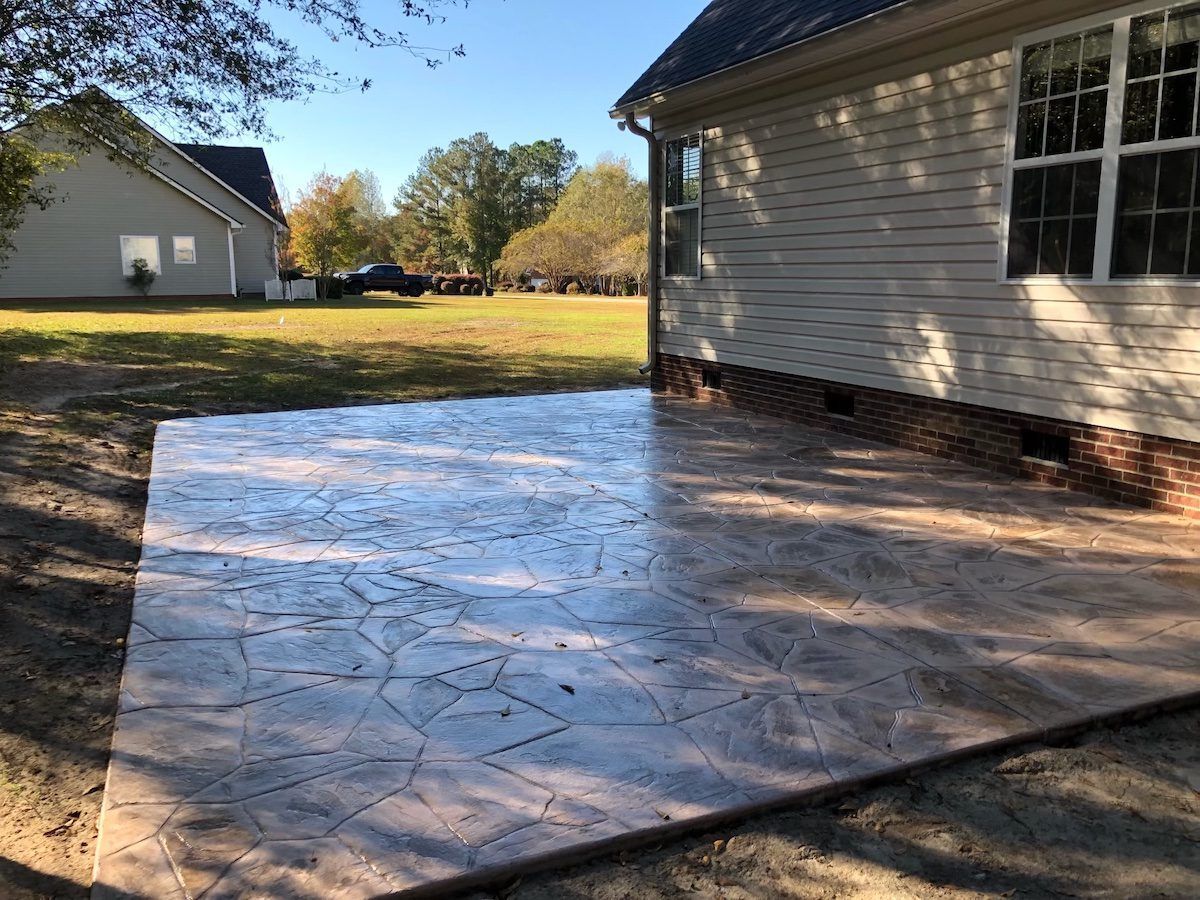 A newly poured stamped concrete patio in a backyard beside a light-sided house under a clear blue sky.