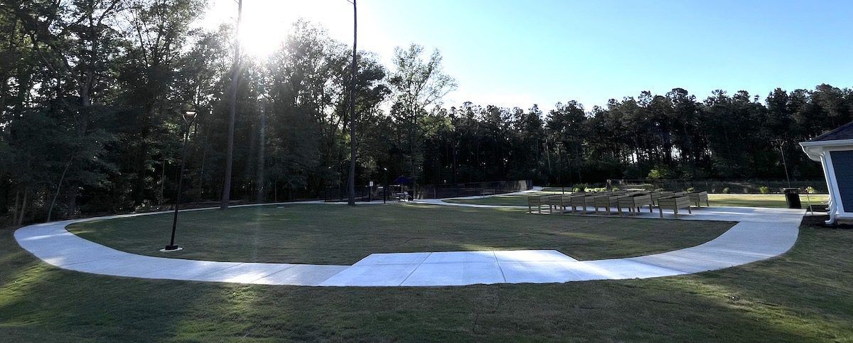 A curved concrete walkway cuts through a grassy lawn toward a tree line under a bright sun, with seating in the distance.