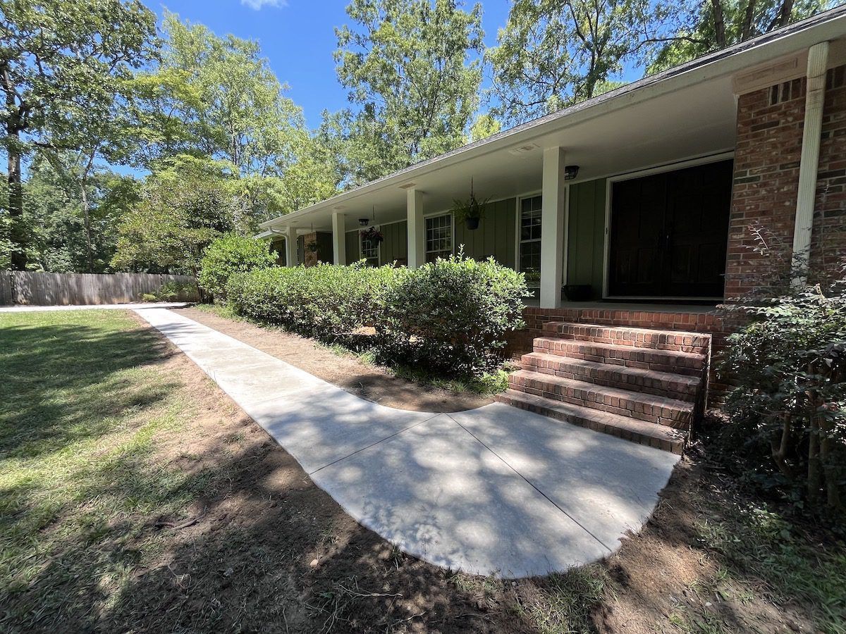 A sidewalk leads to the brick front steps and covered porch of a house surrounded by trees on a sunny day.