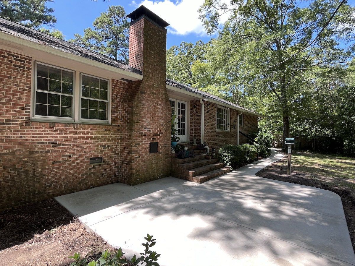 A side view of a red brick house with a brick chimney, a small concrete patio, and steps leading to a side door.