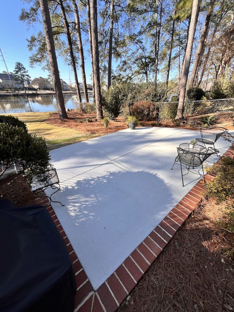 A rectangular patio with a light gray center and red brick border, situated outdoors overlooking a lake and pine trees.