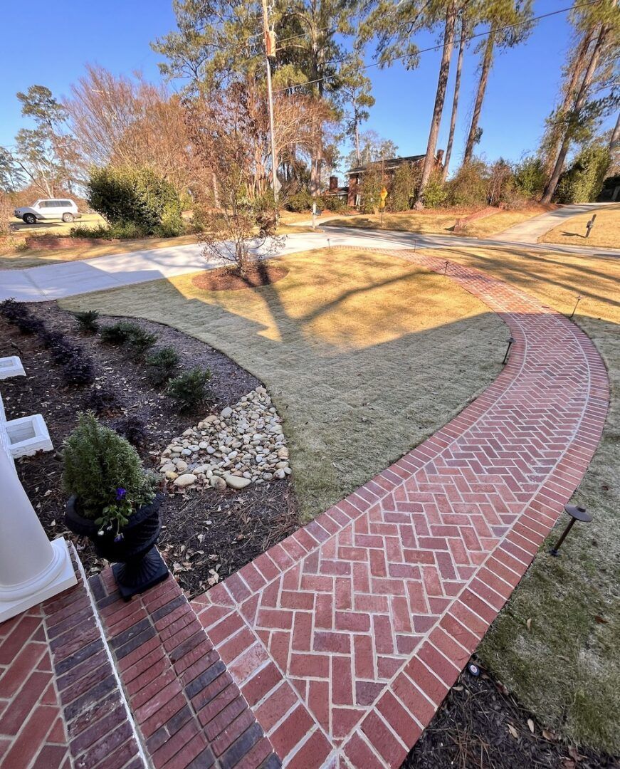 A curved brick walkway leads through a landscaped yard with a lawn, shrubs, and trees under a clear blue sky.