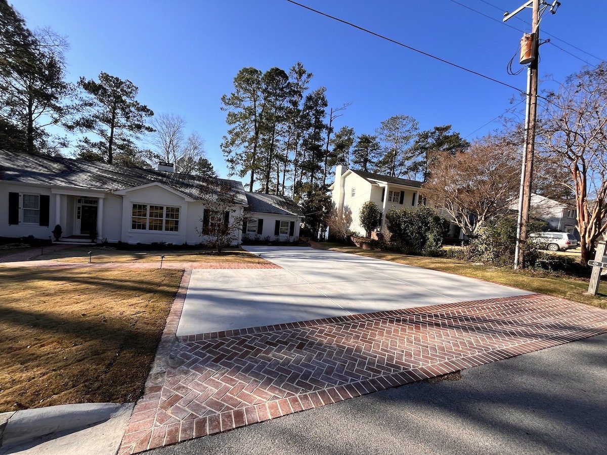 A house with a newly poured concrete driveway and a brick-paved apron set against a sunny, tree-lined suburban street.
