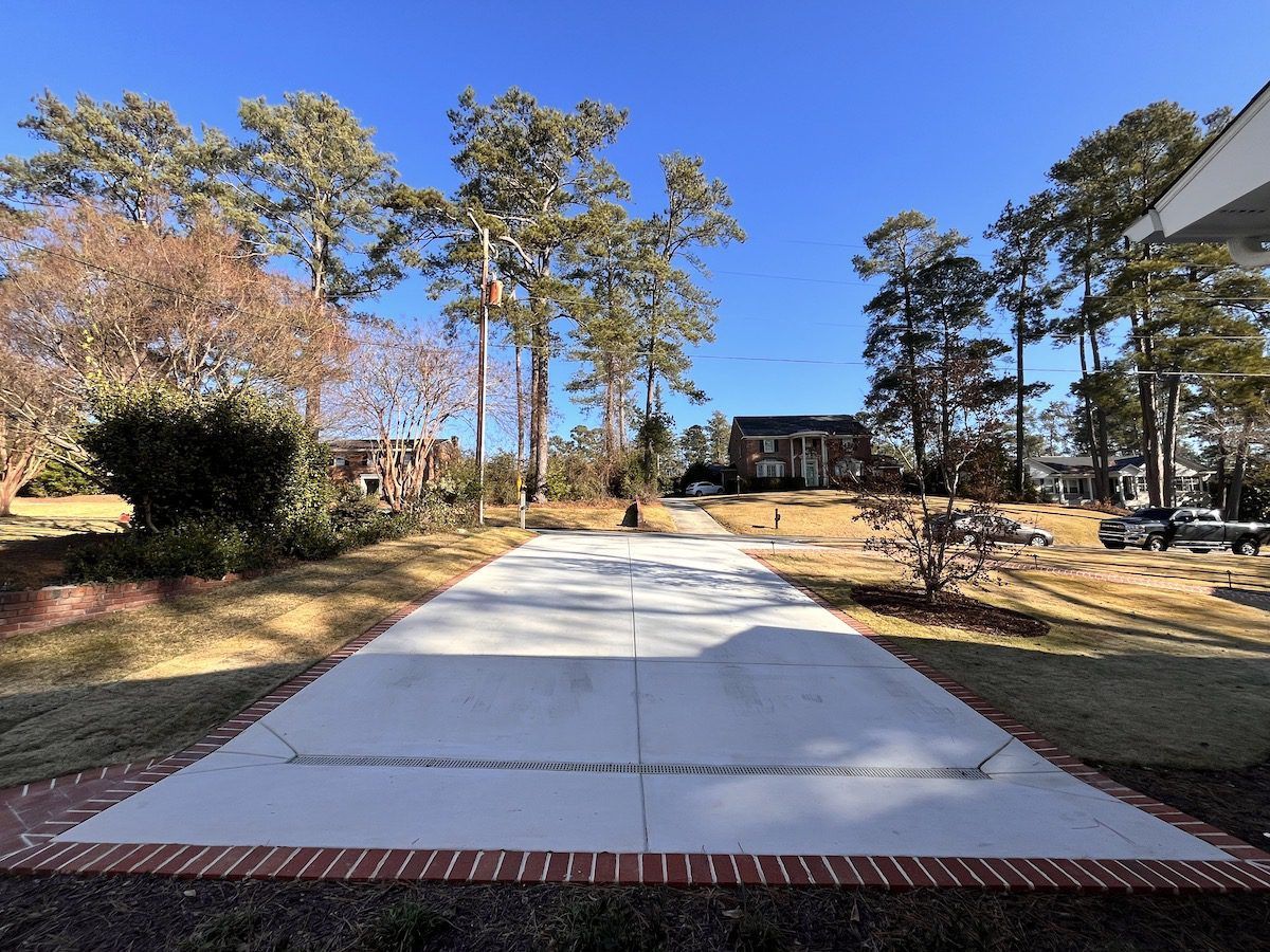 A new concrete driveway with a red brick border leads toward a house surrounded by tall pine trees under a clear blue sky.