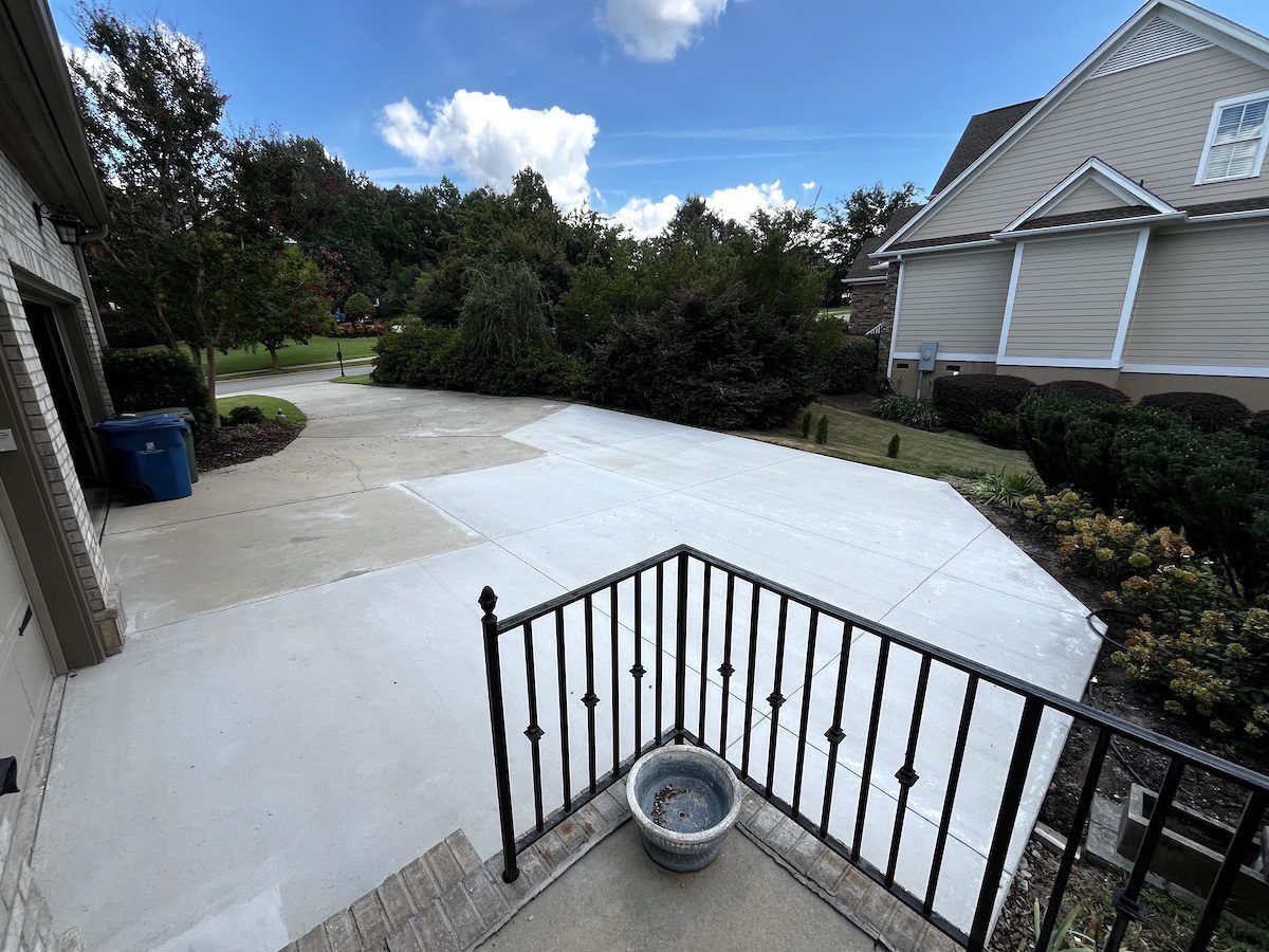 A bright concrete driveway and patio area viewed from an elevated porch featuring a black metal railing and a potted plant.