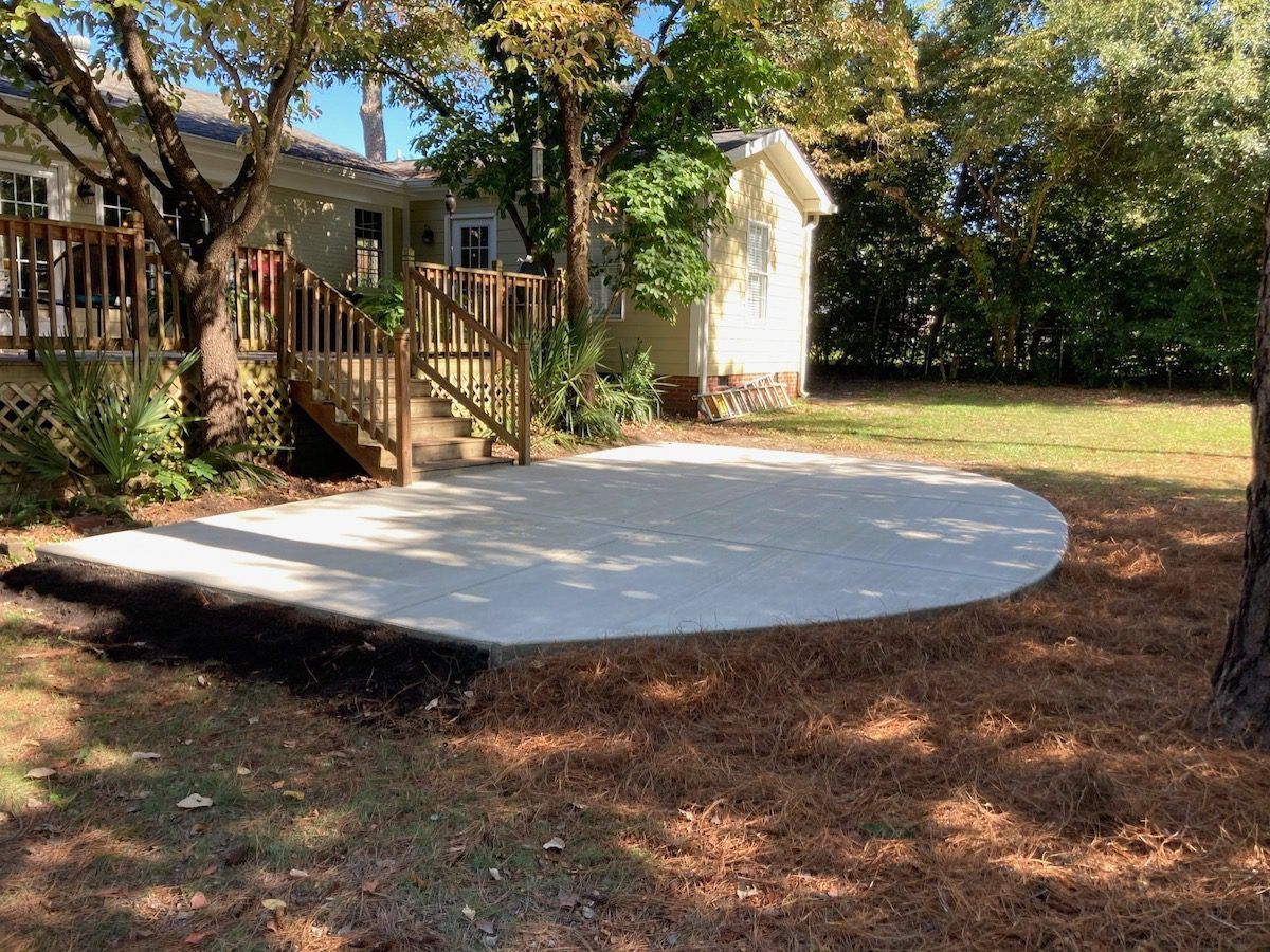 A newly poured, semi-circular concrete patio sits in a backyard next to a wooden deck and house.