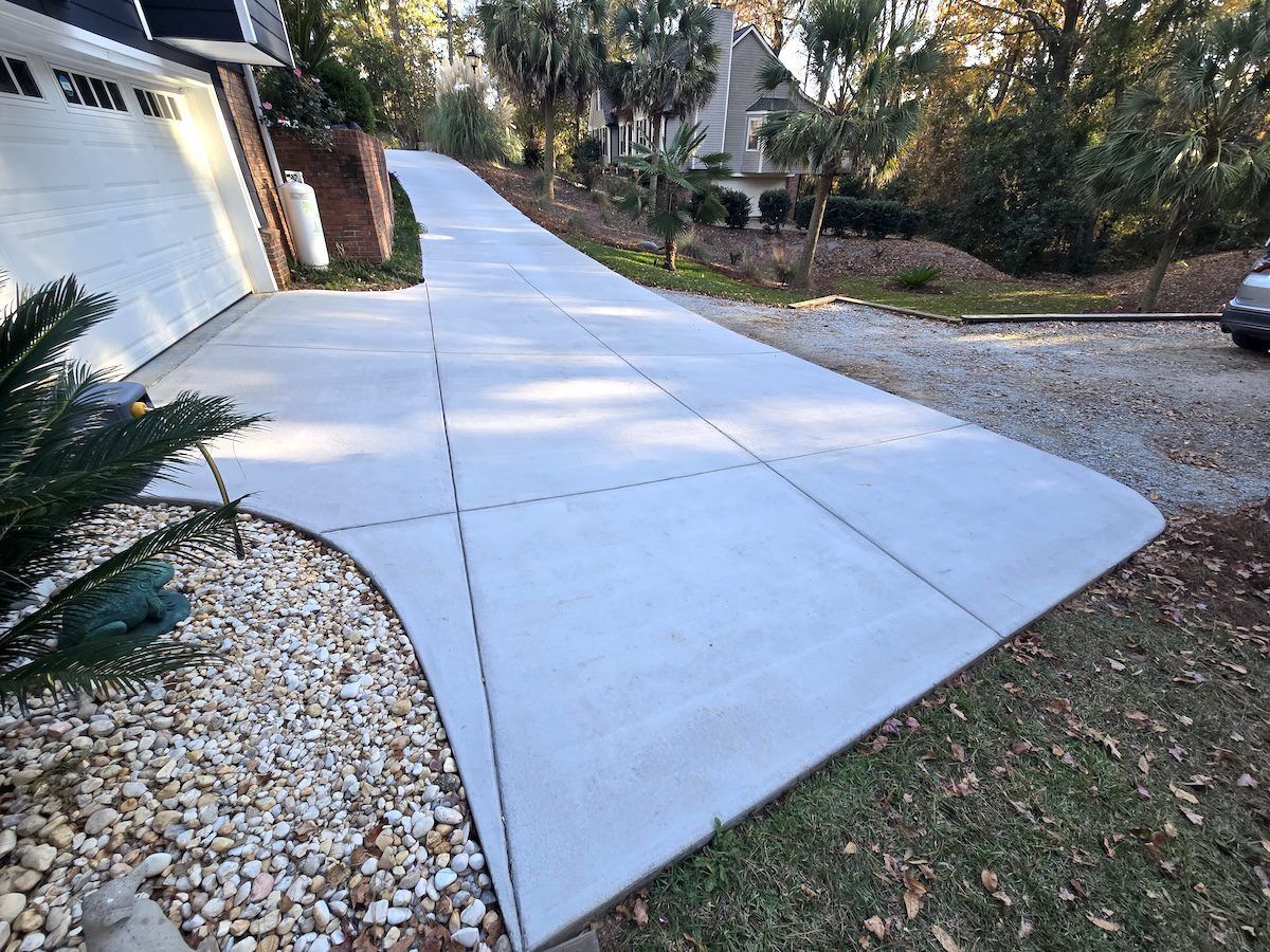 A wide, light-colored concrete driveway with visible expansion joints leading from a garage toward a grassy, wooded yard.