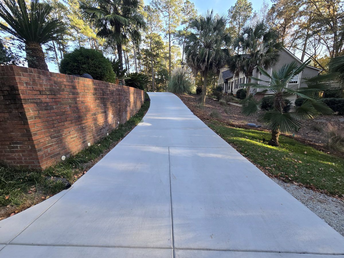 A long, straight concrete driveway leads upward toward a house, flanked by a tall brick retaining wall and trees.