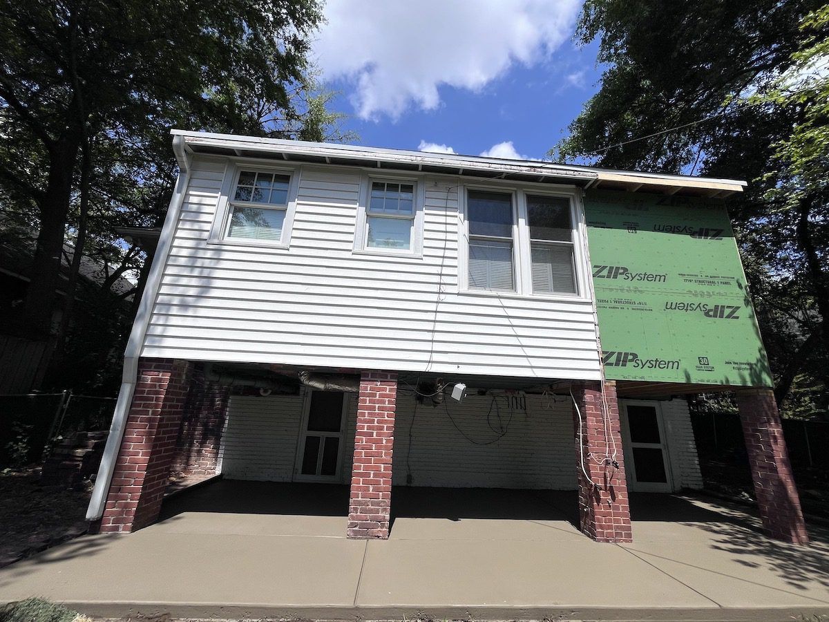An elevated two-story house with white siding and brick support pillars, featuring an unfinished green section on the side.