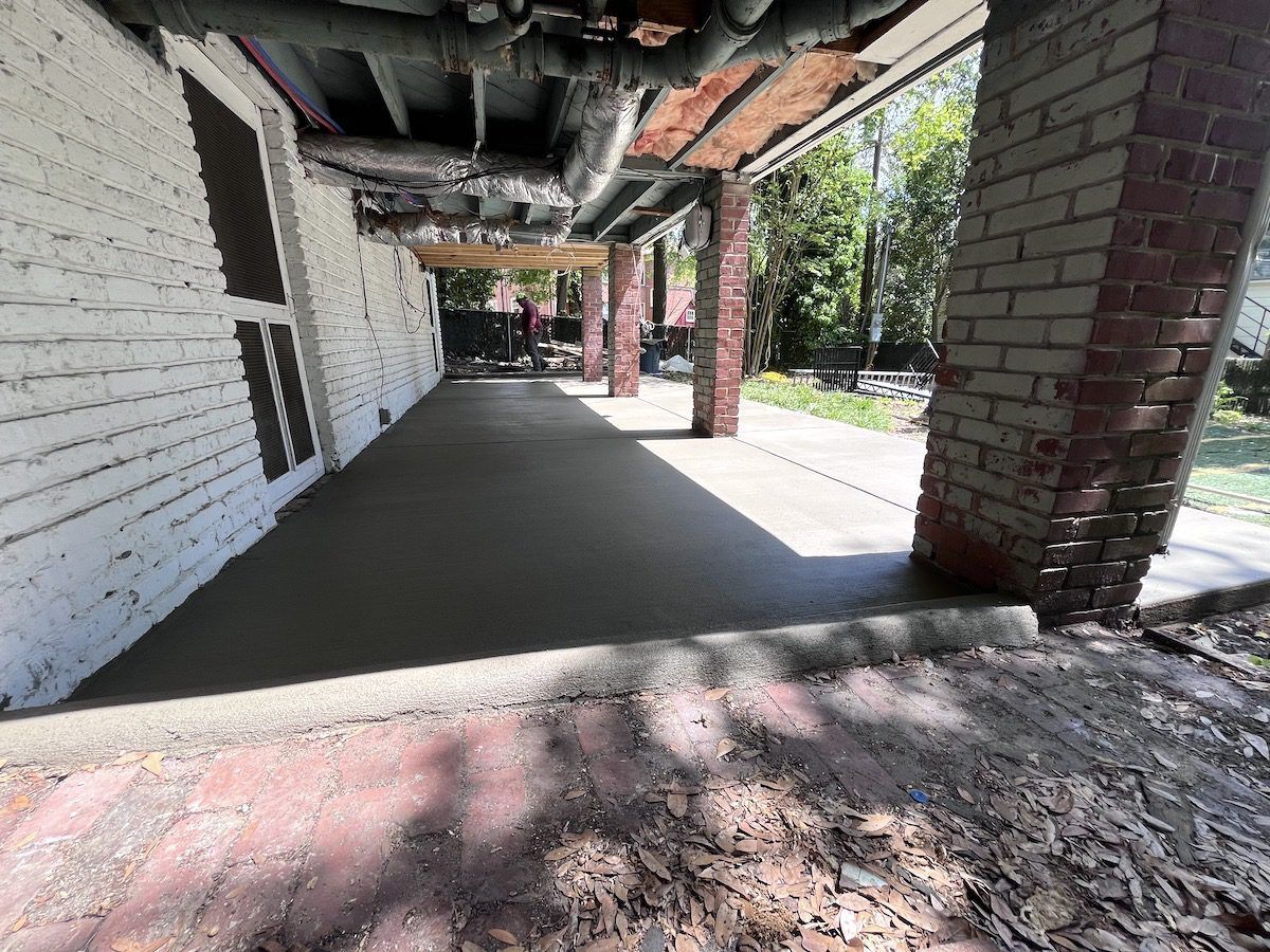 A newly poured concrete patio floor under a covered porch area with a white brick wall and red brick support pillars.