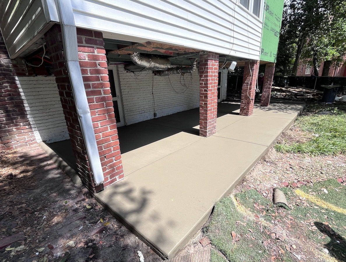 A newly poured concrete patio beneath the elevated, brick-pillared section of a house.