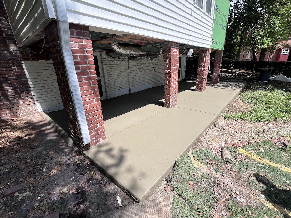 A newly poured concrete patio beneath the elevated, brick-columned section of a house in a residential yard.